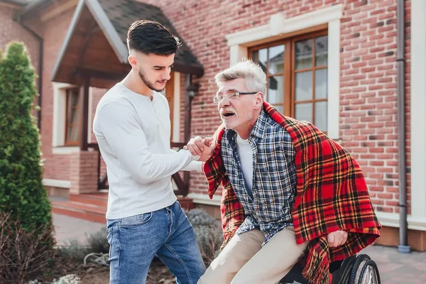 A young man helping an older man with a blanket in front of a brick house