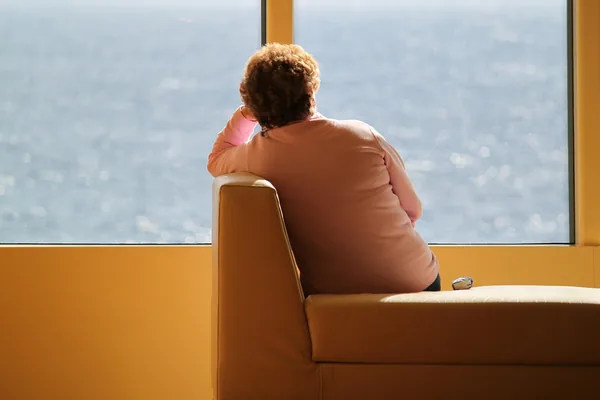 An older person sitting in an armchair looking out a large window