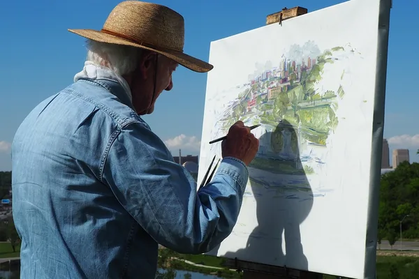 An older man in a straw hat painting a city landscape on a canvas outdoors on a sunny day