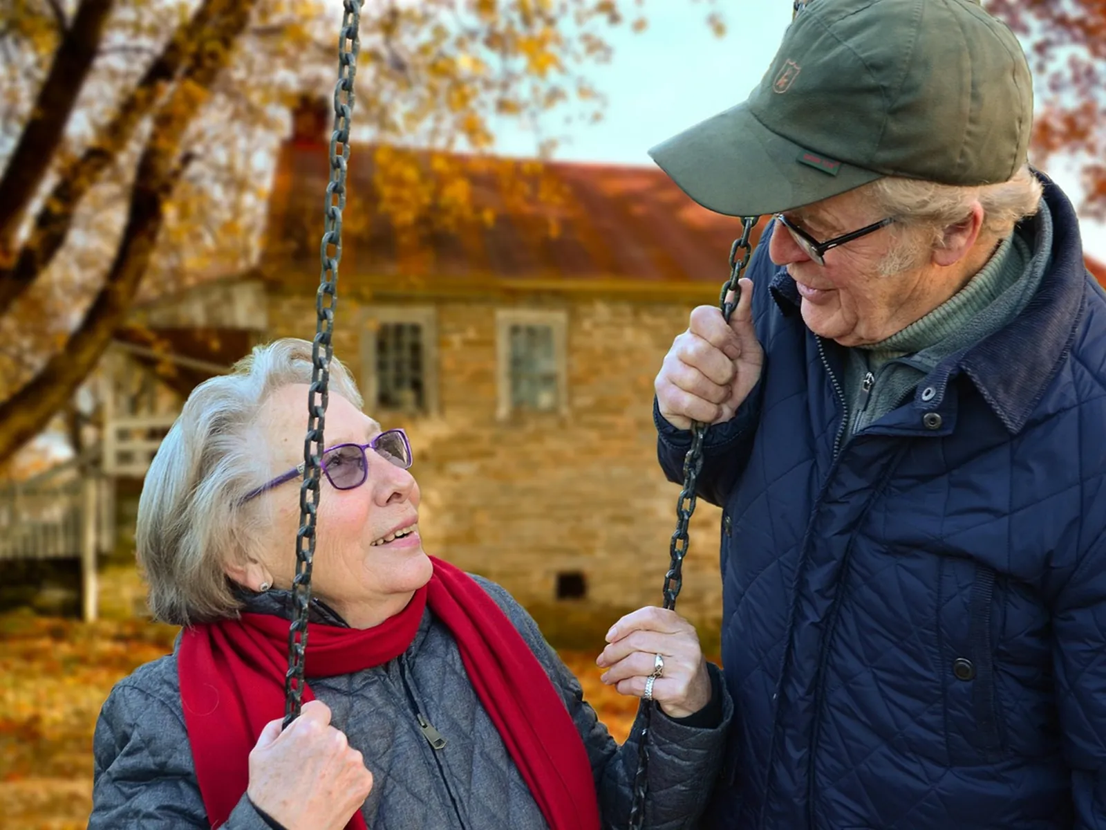 An older woman in a red scarf laughs on a garden swing while her husband in a cap pushes her, with autumn trees behind