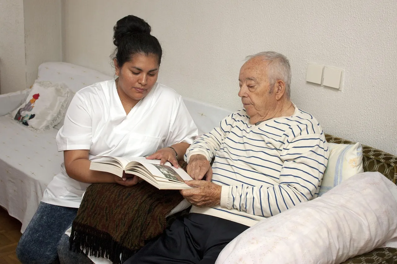 A caregiver sitting beside an older man on a sofa, reading through papers together